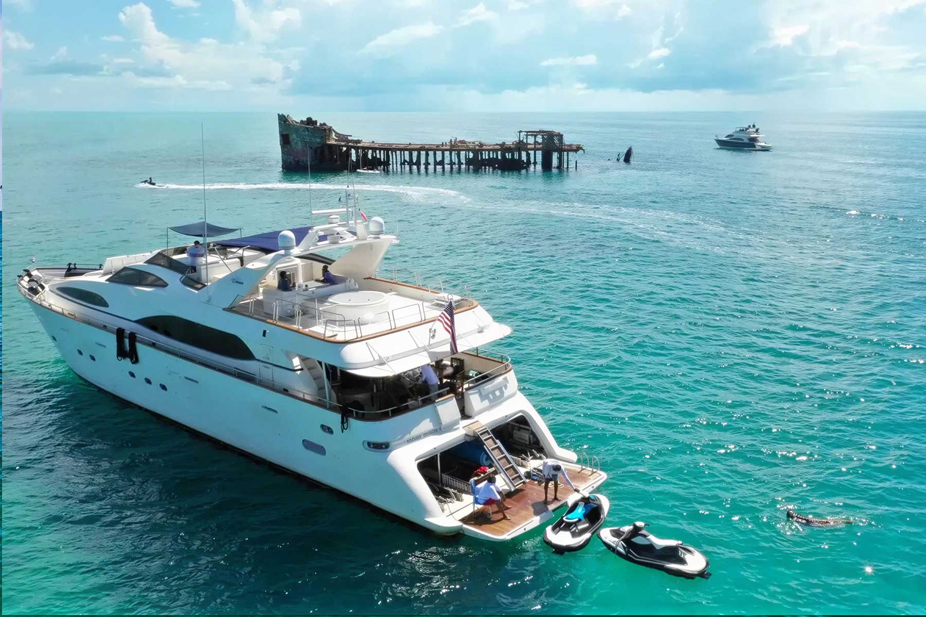 A large white yacht is anchored near an old, rusted shipwreck in clear turquoise water, with a smaller yacht and jet skis nearby under a partly cloudy sky.