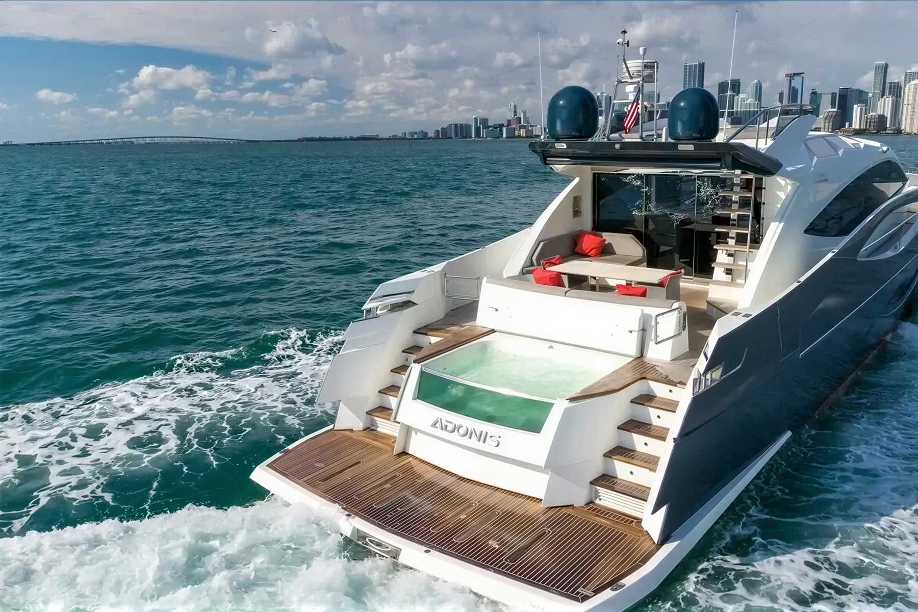 A luxury yacht named "Adonis" cruises on blue water near a city skyline, featuring a deck with a hot tub, seating area, and two staircases.