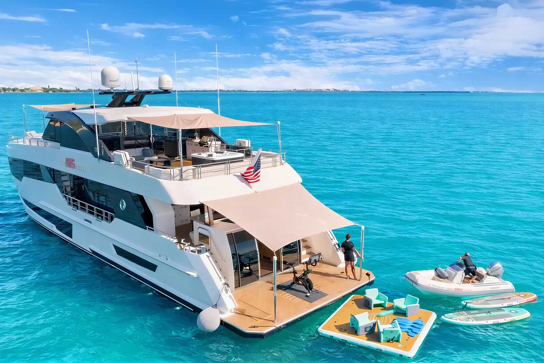 A large white yacht anchored in clear blue water with a floating lounge, jet ski, paddleboards, and two people enjoying water activities.