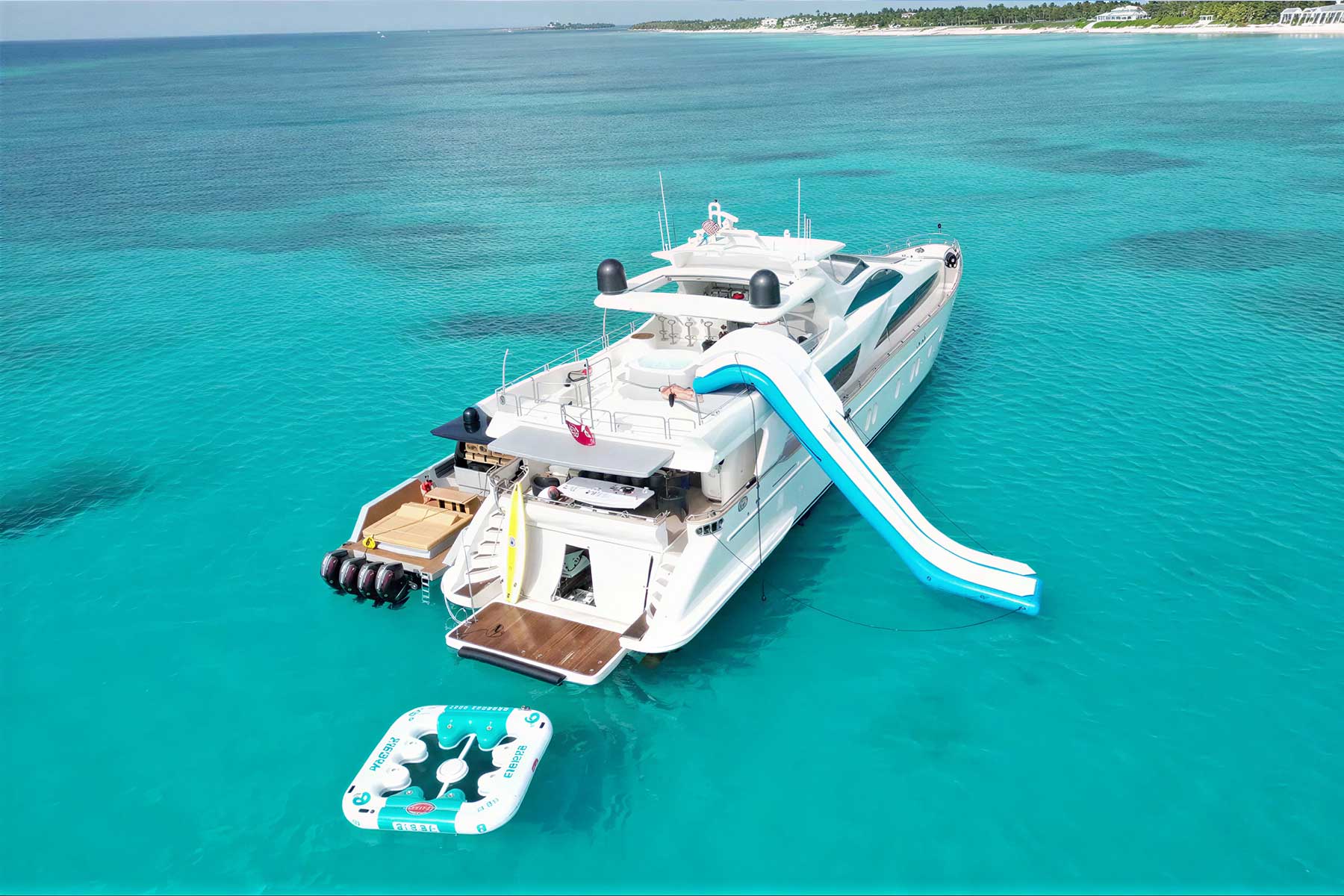 A white luxury yacht with a blue and white slide is anchored in clear turquoise water, with an inflatable float attached at the back.