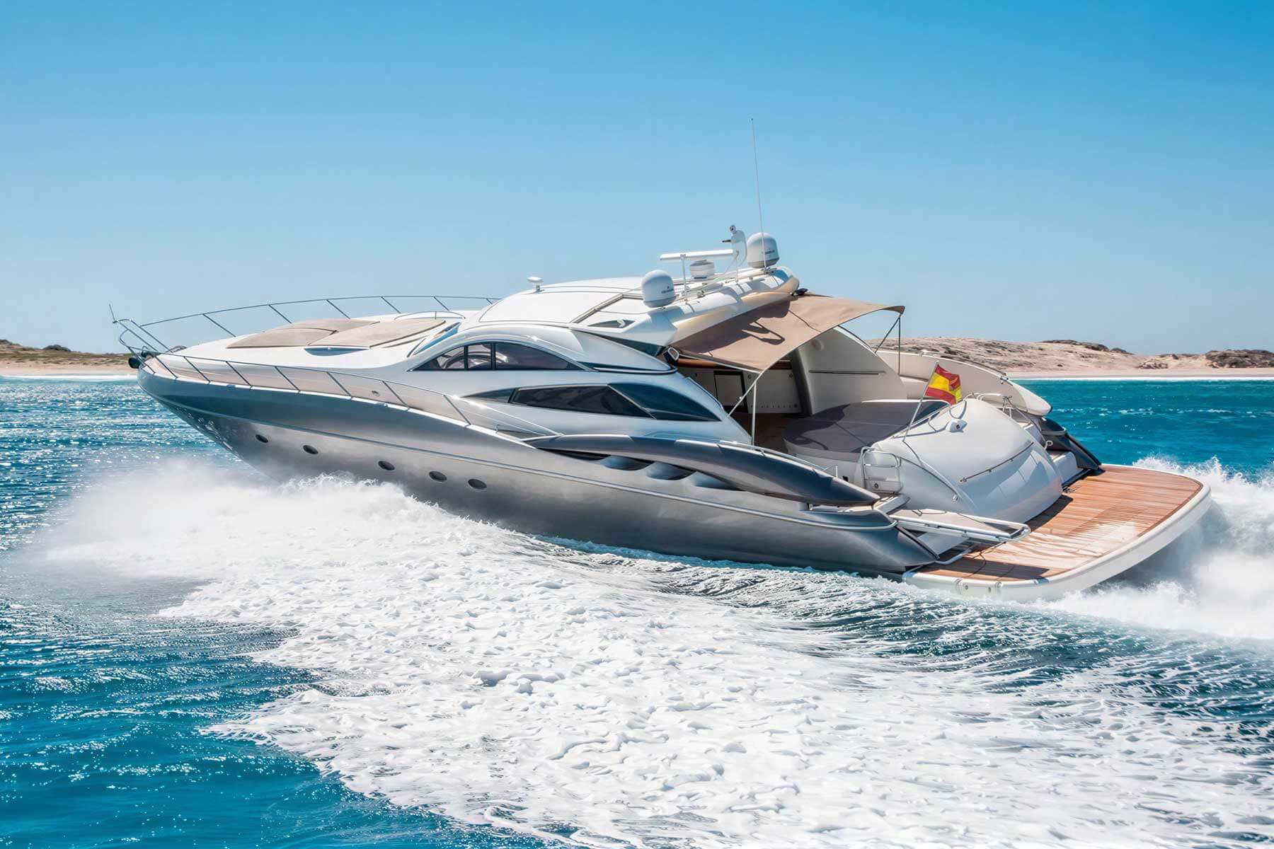A sleek, modern yacht cruises quickly across blue water near a sandy shoreline under a clear sky.