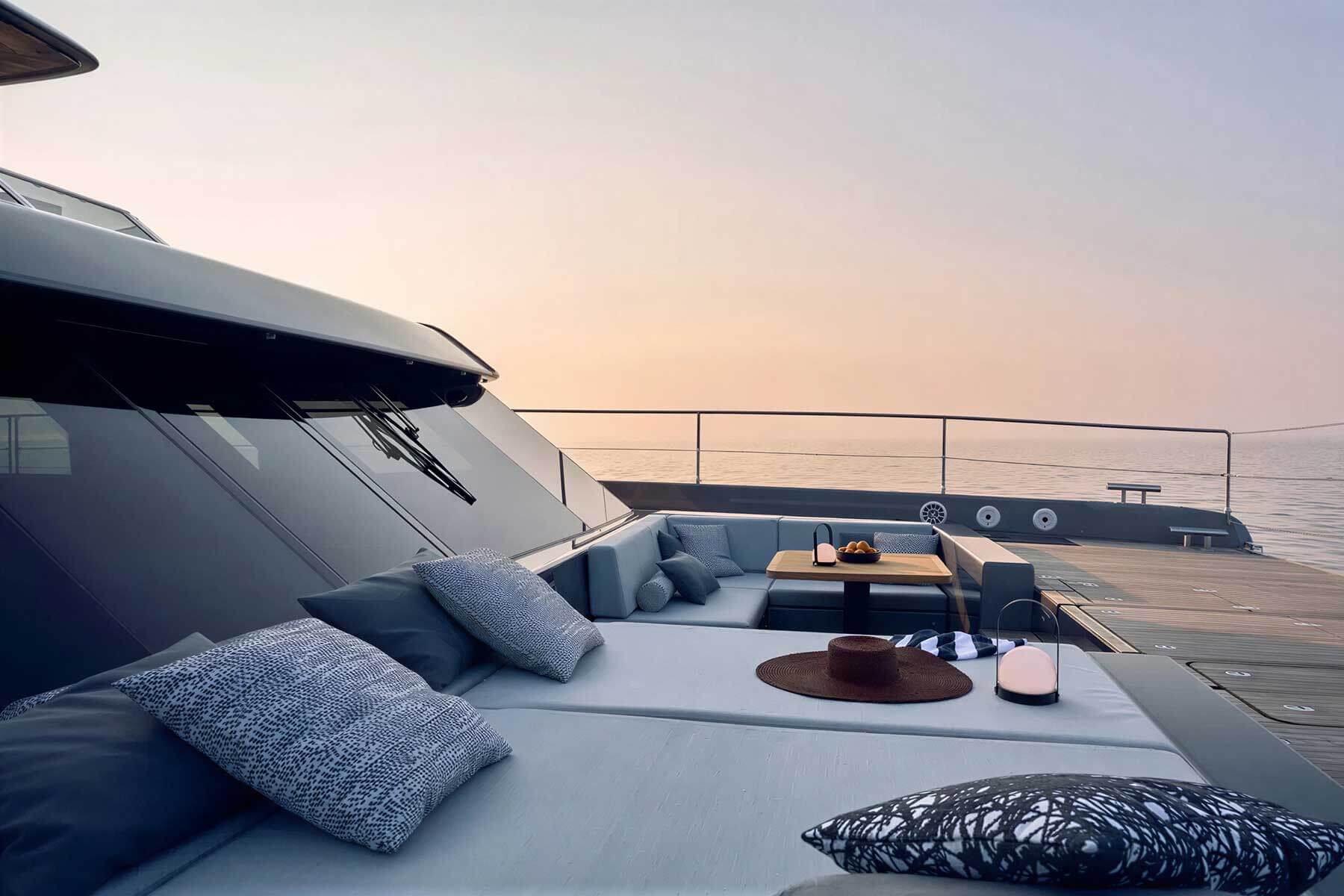 Modern yacht deck with cushioned seating, decorative pillows, a table with fruit, a hat, and lanterns, overlooking calm sea at sunset.
