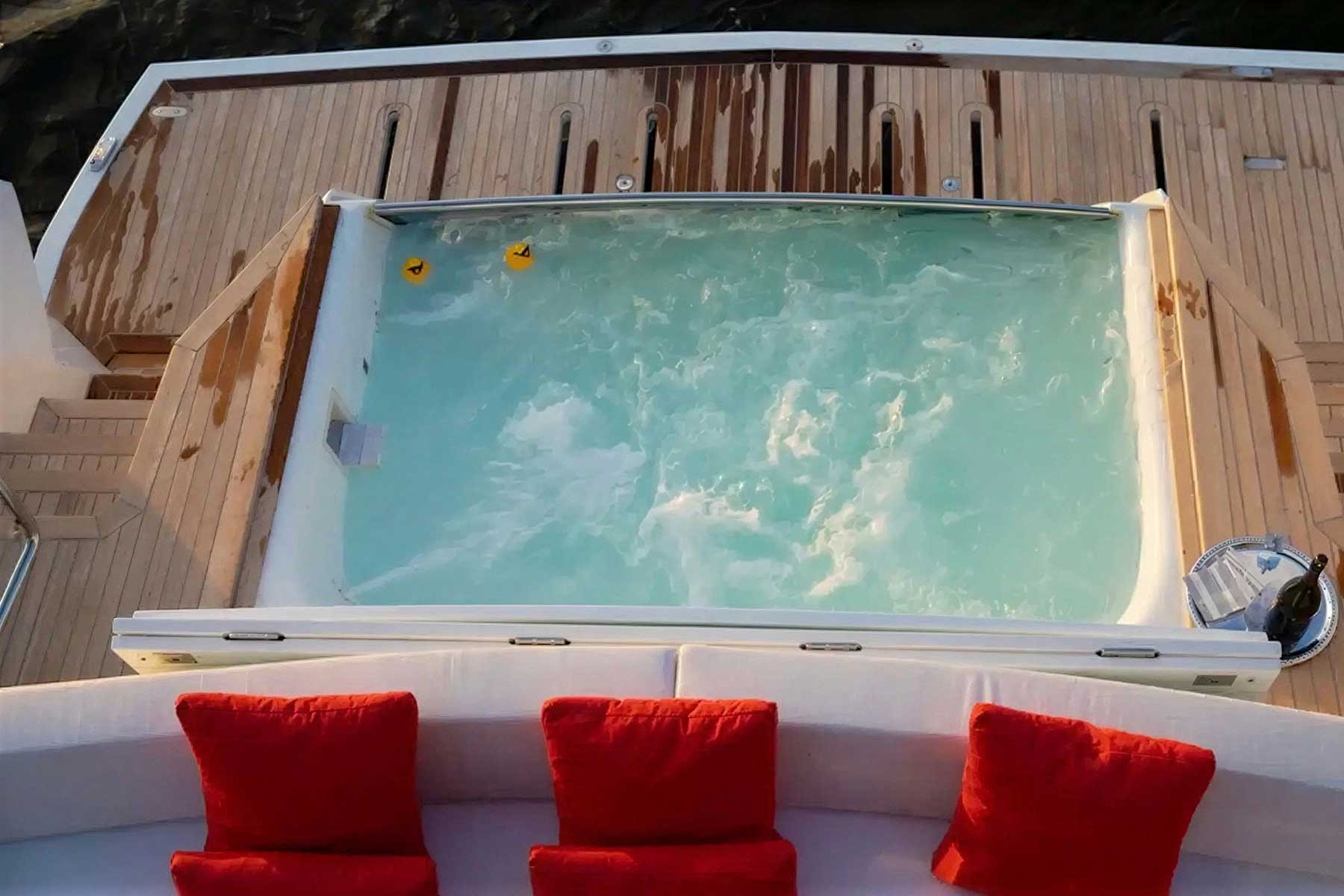 A view from above of a hot tub with swirling water on a yacht deck, with three red cushions on a seating area in the foreground and two rubber ducks floating in the tub.