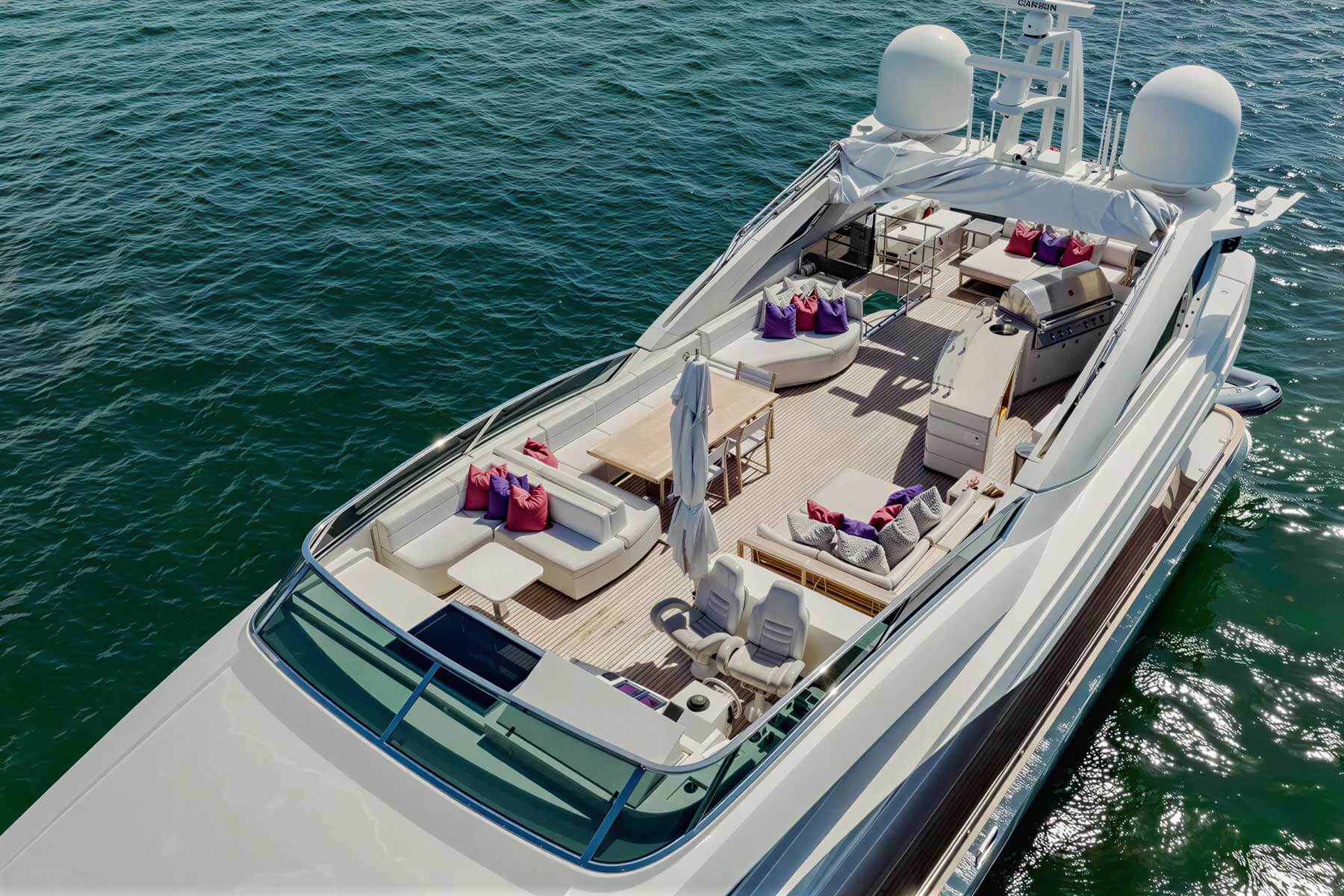 A luxury yacht with spacious outdoor seating, tables, and modern amenities is docked on calm water under sunny conditions.