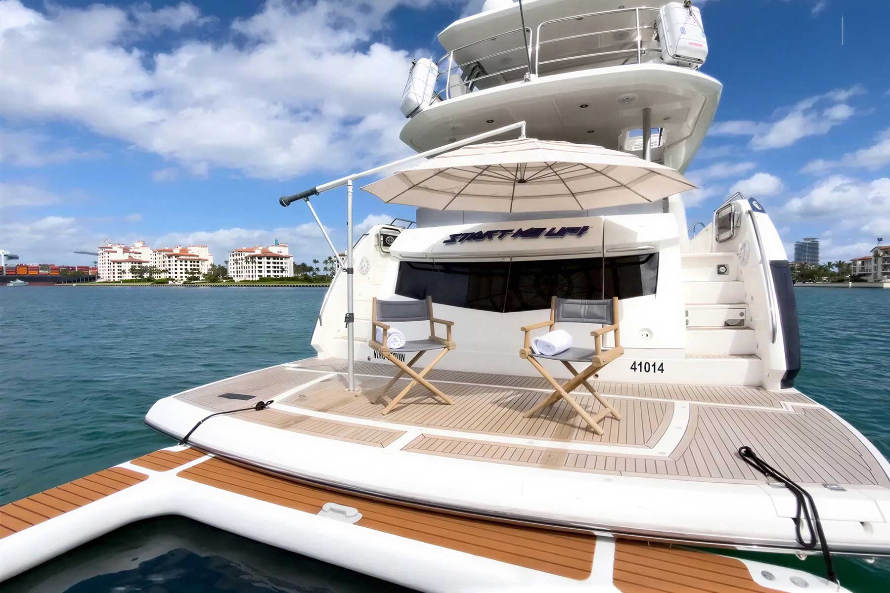 Yacht rear deck with two chairs and an umbrella facing the water; waterfront buildings visible in the background under a partly cloudy sky.