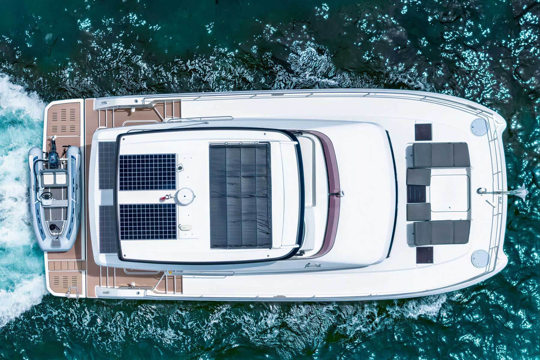 Aerial view of a white yacht with solar panels on the roof, a lounge seating area, and a small boat attached to the stern, cruising through blue-green water.