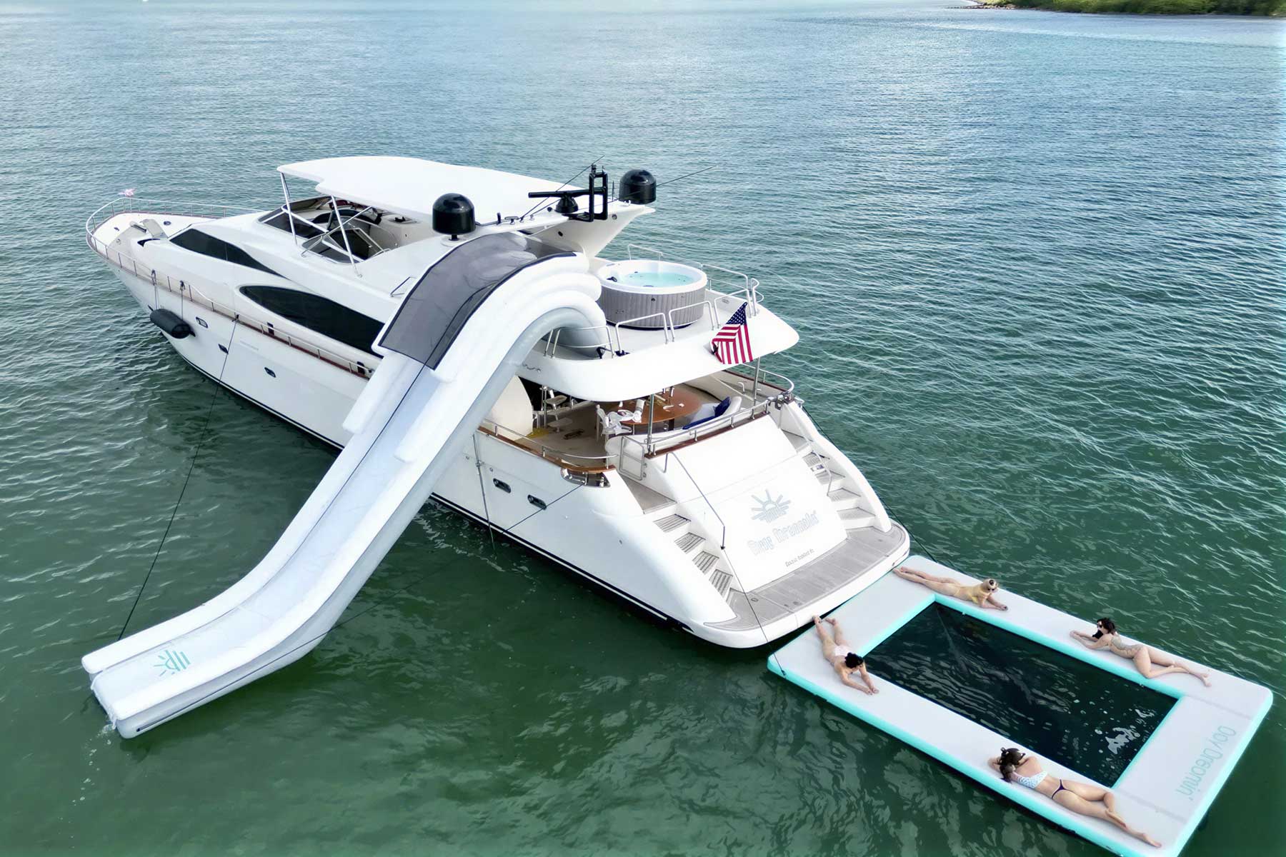 A large white yacht floats on calm water, featuring a waterslide and an attached floating pool with four people relaxing.
