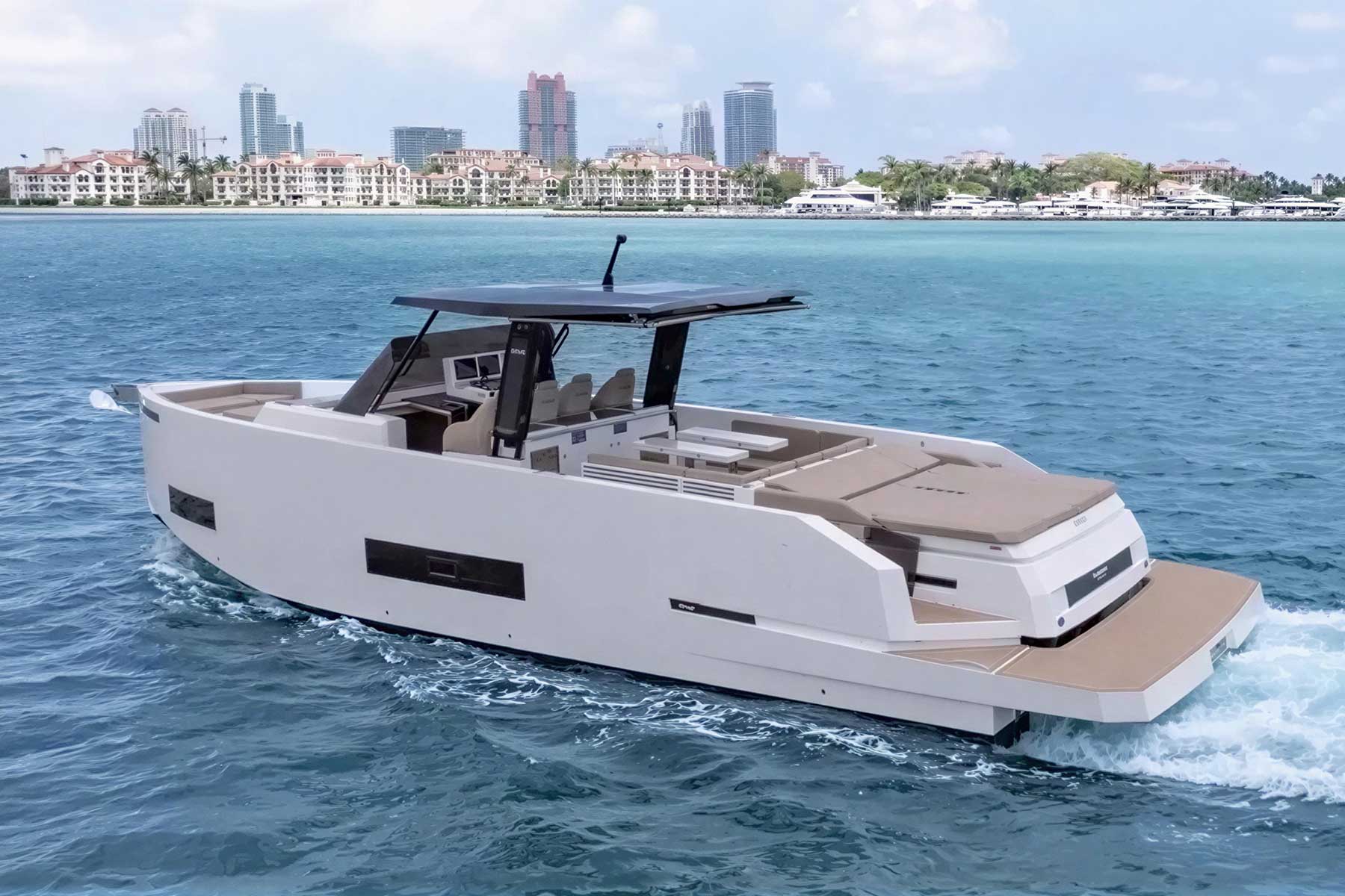 A modern, white motor yacht cruises on blue water with a city skyline and waterfront buildings in the background under a partly cloudy sky.