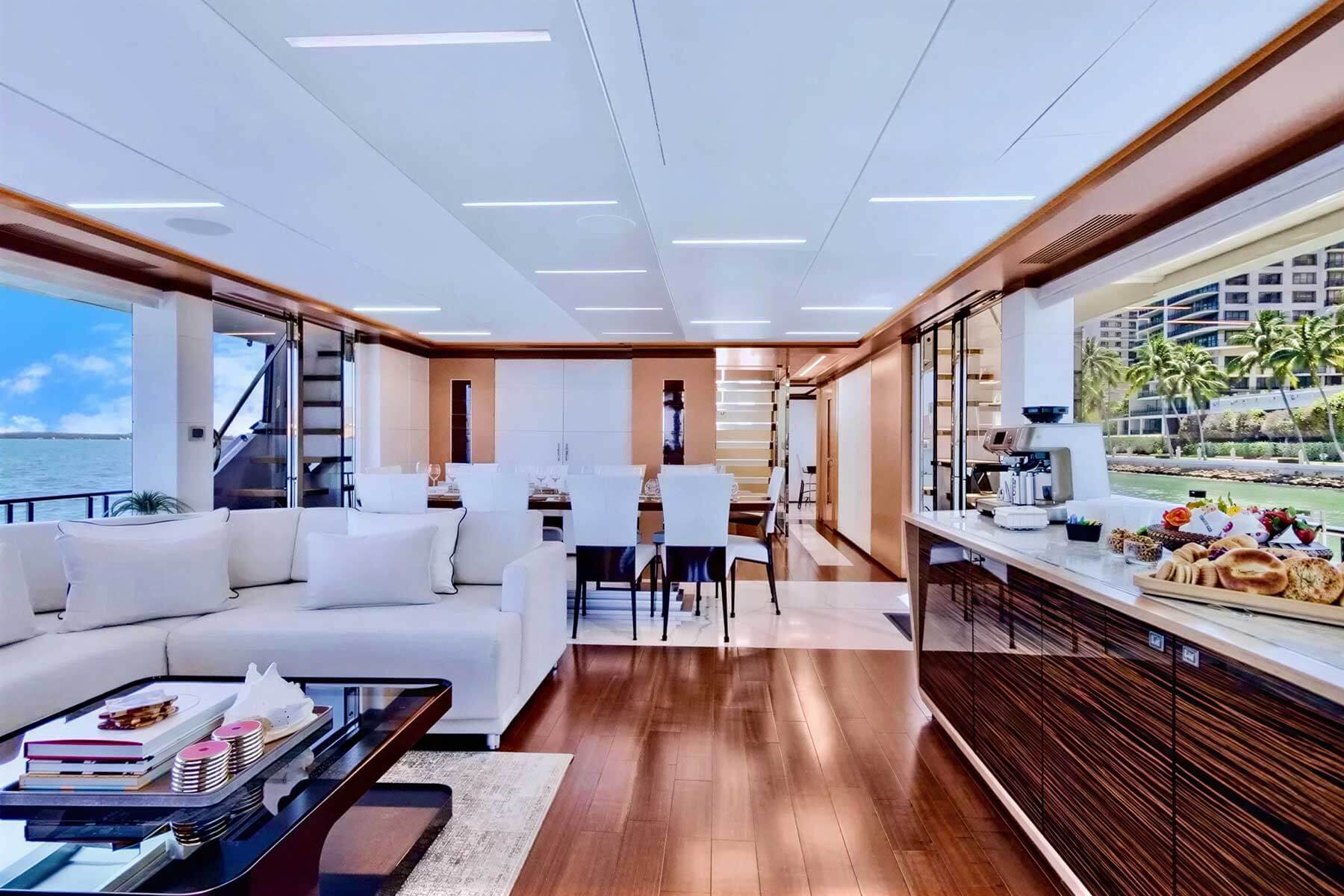Modern yacht interior with white sofas, dining table with chairs, wooden floors, large windows showing water and cityscape, and a side counter set with food and drinks.