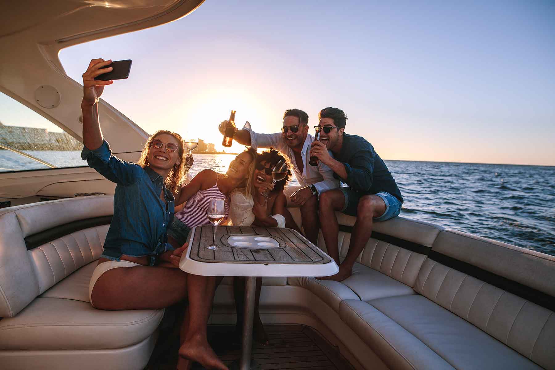 group-photo Five people sit together on a boat at sunset, taking a group selfie and holding drinks, with the ocean visible in the background.