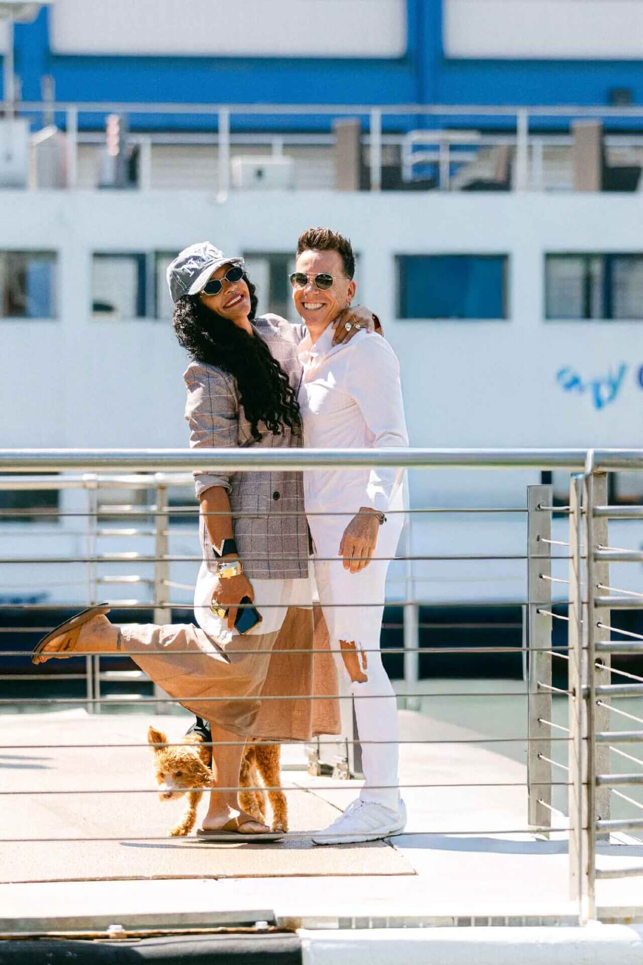 jeff-and-valentina-on-deck Two people stand smiling on a dock, with one person's leg playfully raised and a small dog at their feet. A large boat is visible in the background.
