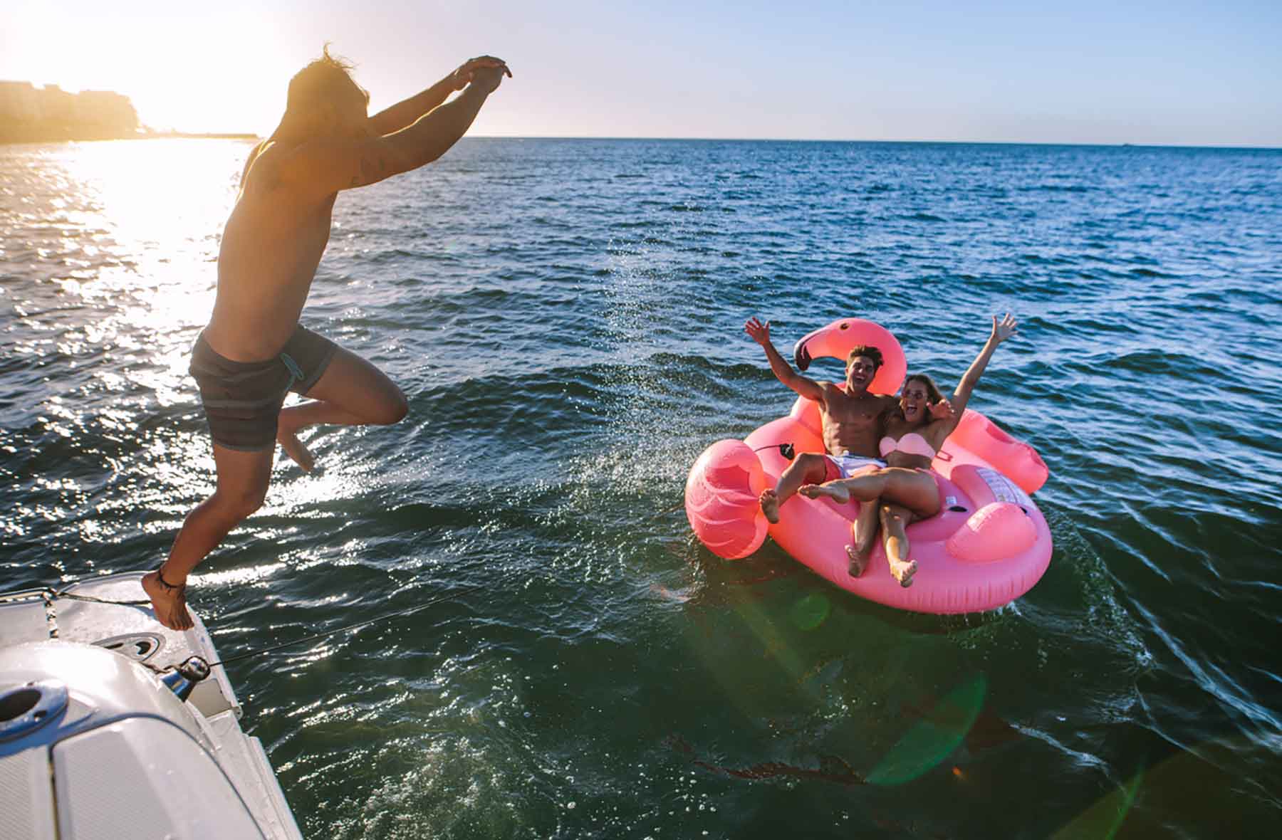 swimming-02 A man jumps off a boat into the ocean while two people relax and wave on a large pink flamingo float nearby.